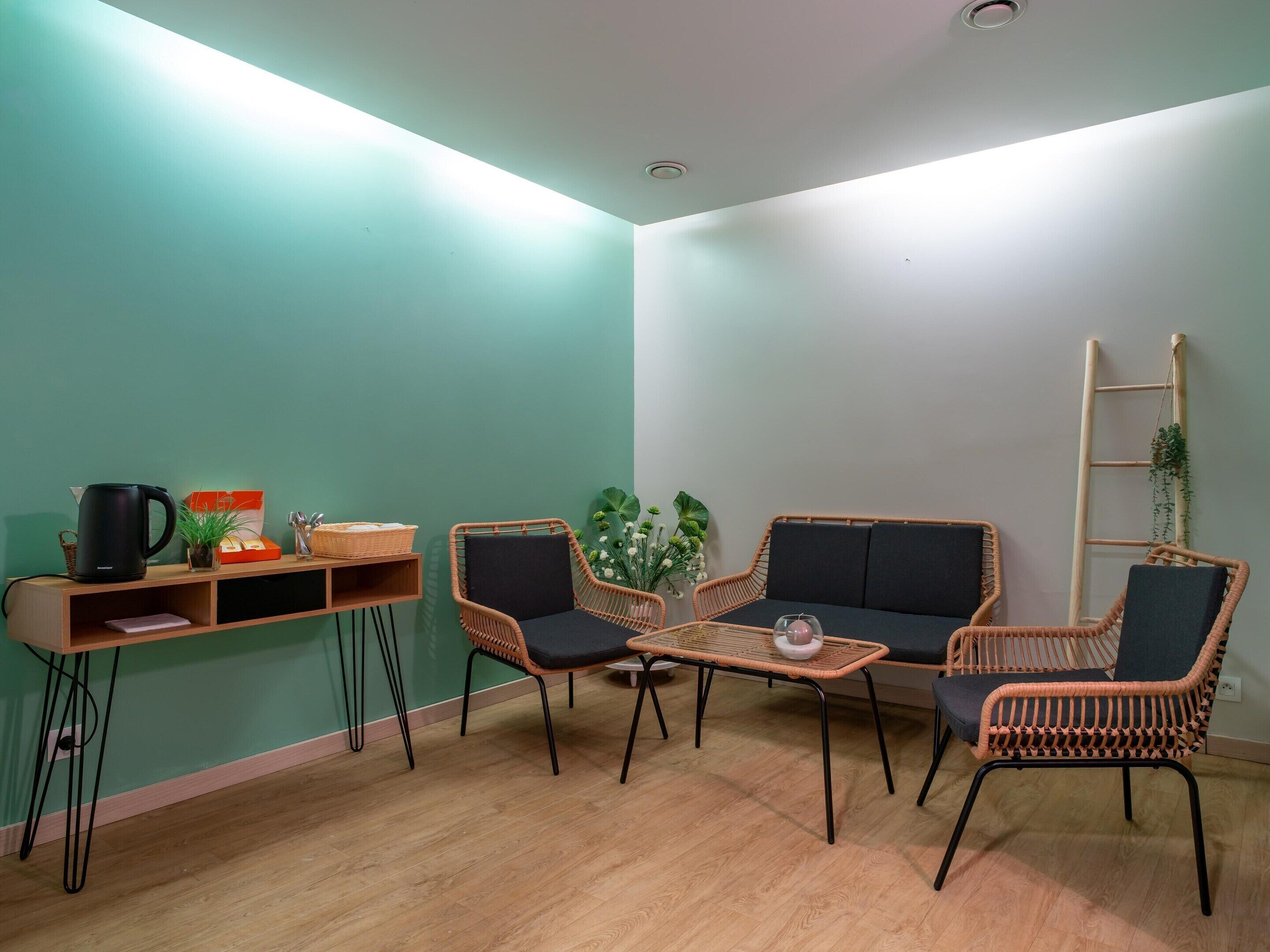 Una habitación con piso de madera, una cafetera negra, sillas de mimbre, una pequeña mesa y una escalera apoyada en la pared.