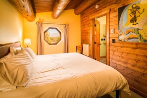 A wooden cabin room with a bed, a window with curtains, a painting on the wall, and a wooden ceiling. - Purple Hobbit Home / Hot Tub / Sauna / Fire Pit (Crestone)