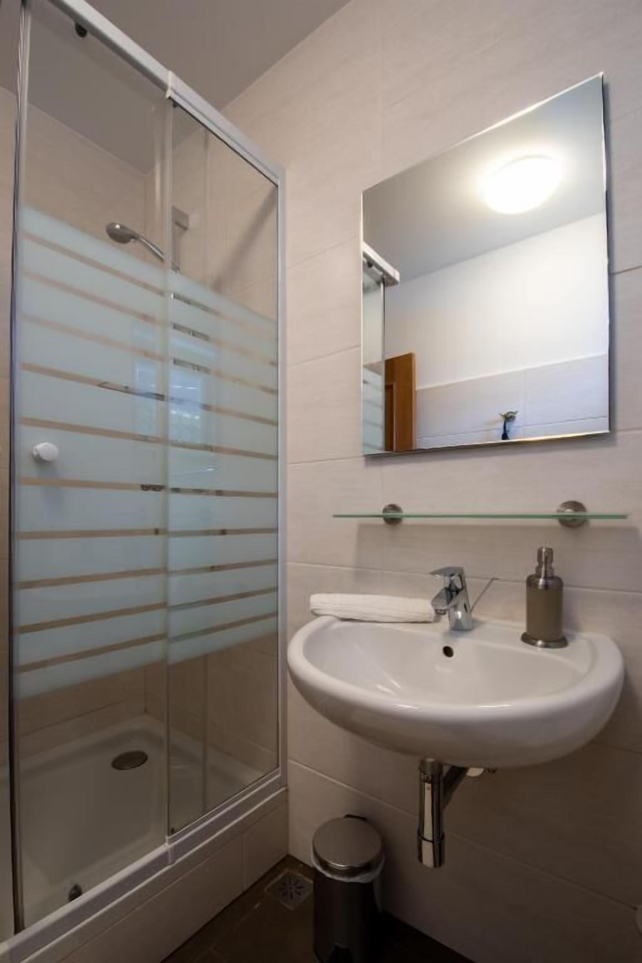 A bathroom with a glass shower enclosure, a white sink with a mirror above it, and a small trash bin.