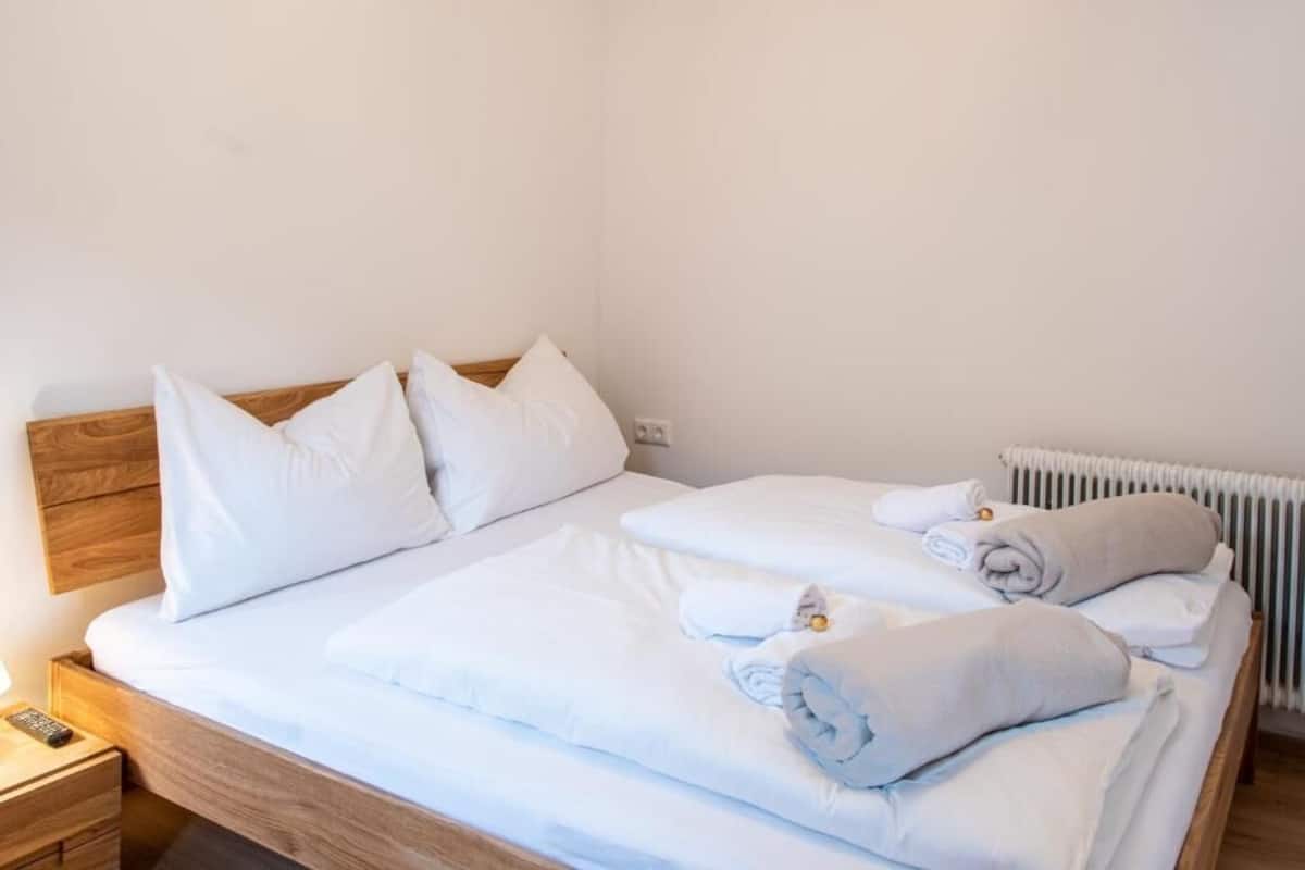 A neatly made bed with white linens and rolled towels, a wooden headboard, a bedside table with a remote control, and a radiator.
