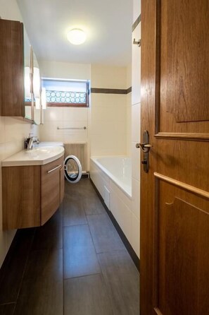 Une salle de bain avec une porte en bois, un lavabo blanc, un miroir, une fenêtre et une baignoire.