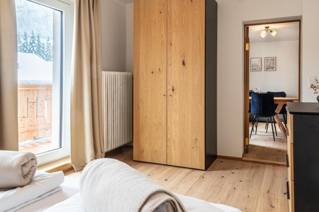 Una stanza con un letto, un armadio in legno, un termosifone, una finestra con tende e un'area pranzo con tavolo e sedie, con vista su alberi.