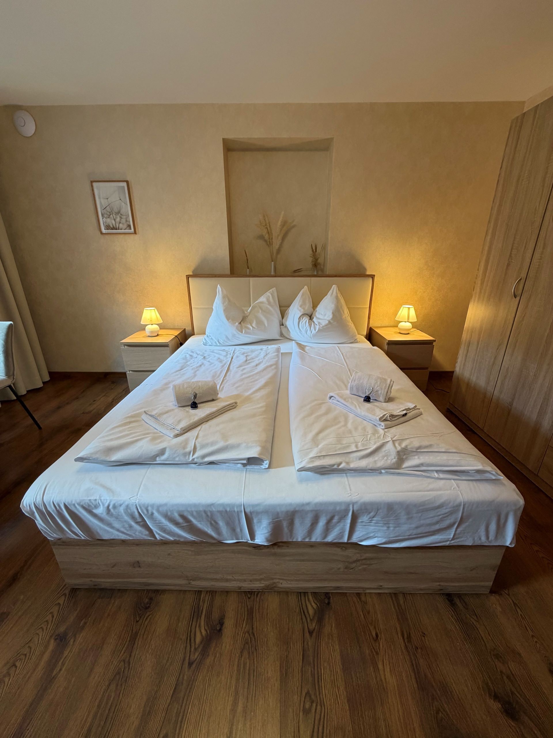 A double bed with white linens, two bedside lamps, a framed artwork on the wall, and a wooden floor.