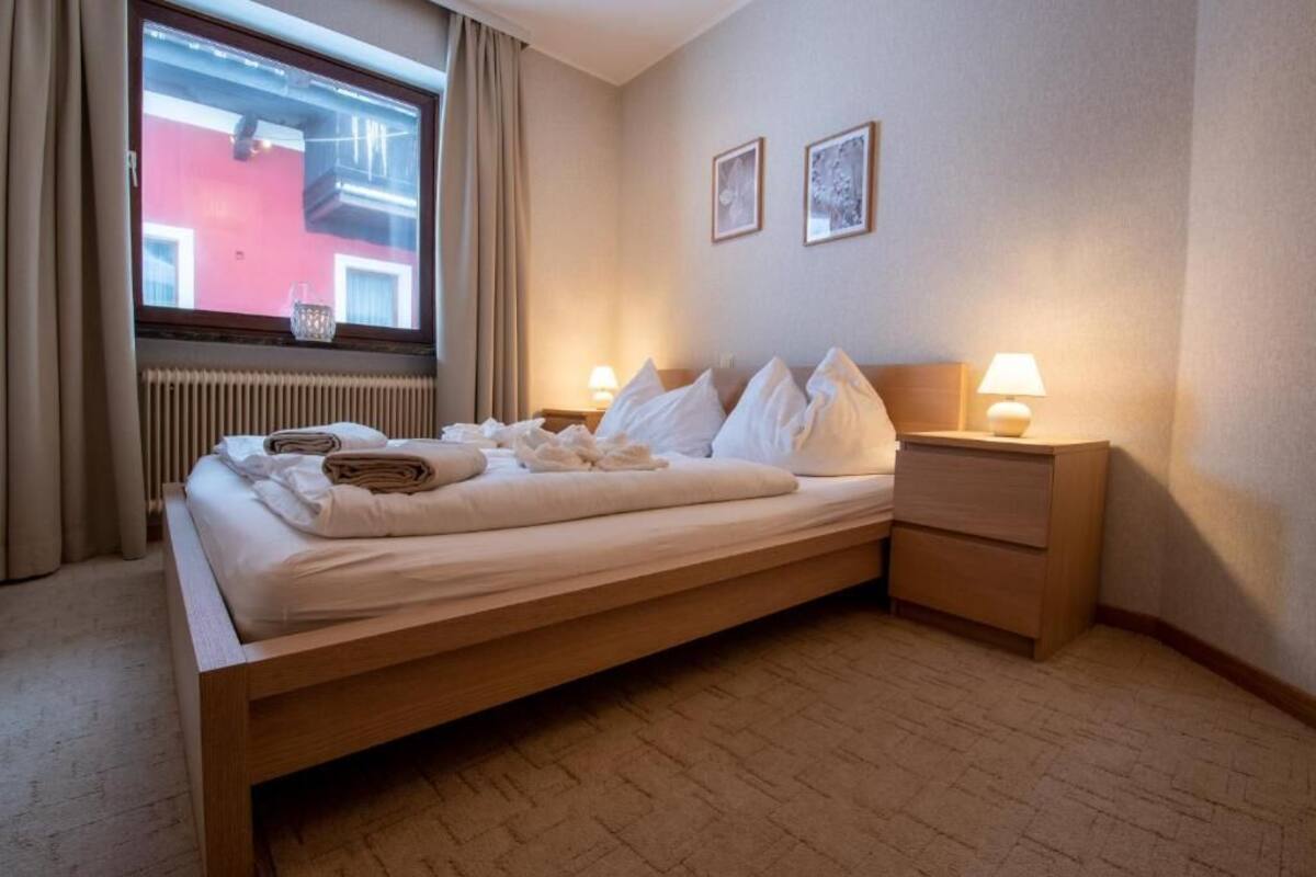 A neatly made bed with pillows and a folded towel, a bedside table with a lamp, a window with curtains, and a radiator.