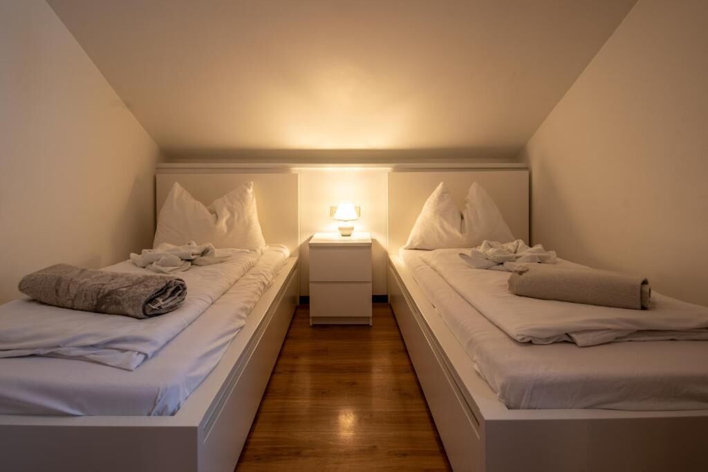 A small, dimly lit room with two single beds, a nightstand with a lamp, and a wooden floor.