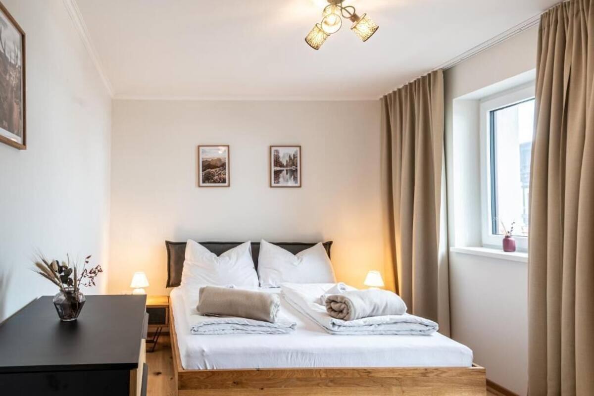 A bedroom with a wooden bed, two bedside lamps, a window with curtains, and two framed pictures on the wall.