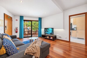 A modern living room with a grey sofa, a television, and a dining area visible through a glass door.