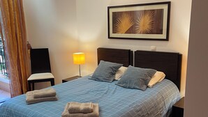 Un letto rifatto con cura, con una coperta blu, due cuscini e due asciugamani piegati. Sopra il letto si trova un’opera d’arte incorniciata e una lampada a lato.