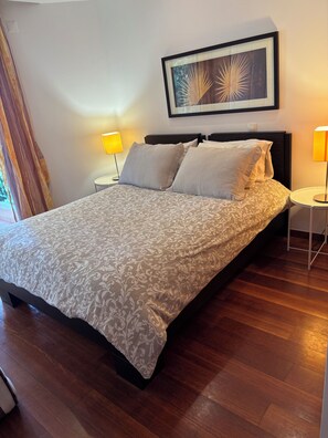 A neatly made bed with a patterned bedspread, two bedside lamps, a framed artwork above the bed, and a wooden floor.