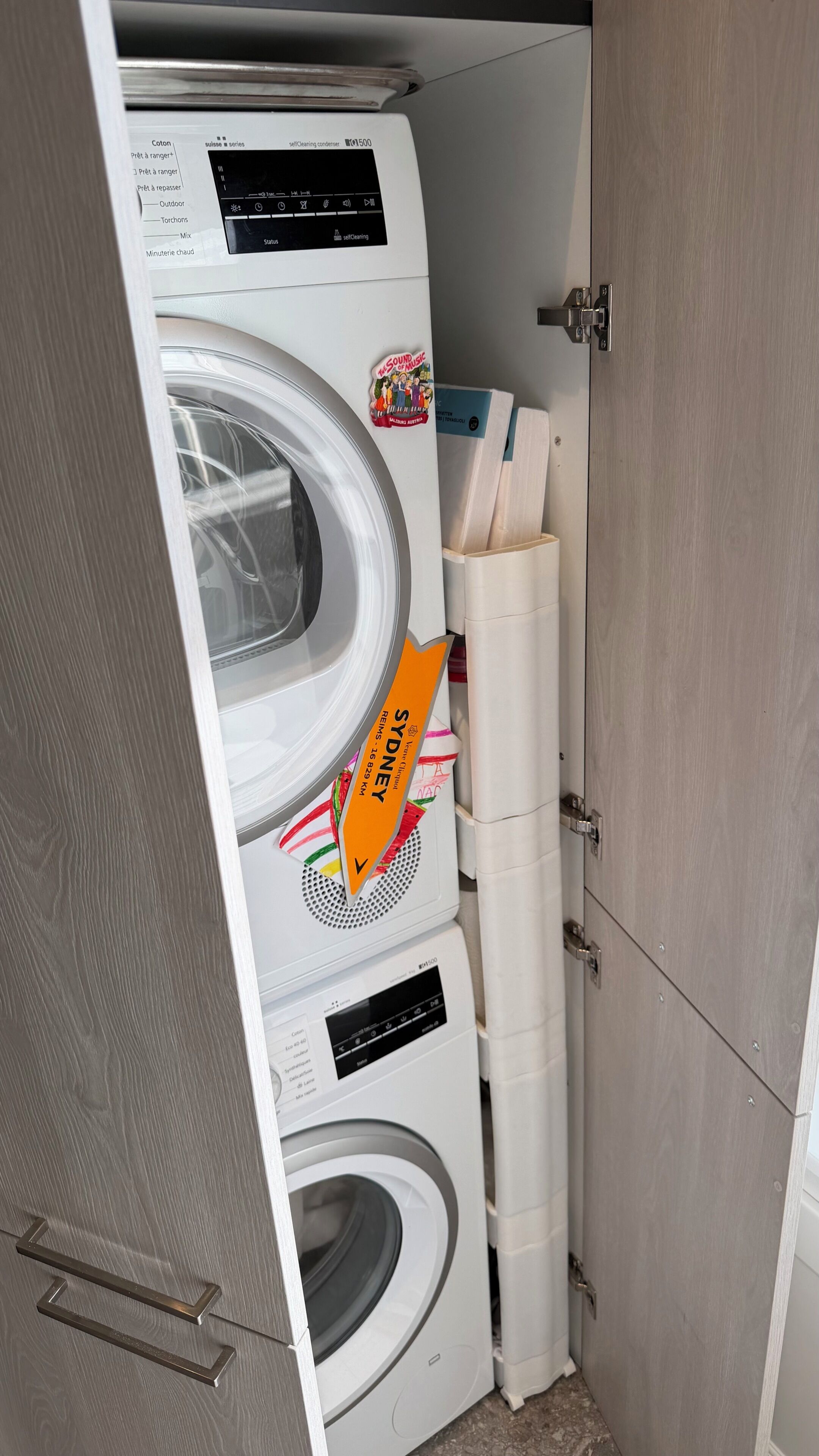 Ein kompakter, moderner Waschraum mit einer Frontlader-Waschmaschine und einem Trockner, einem Holzschrank und einer kleinen Ablage mit einem Hinweis auf den Standort.
