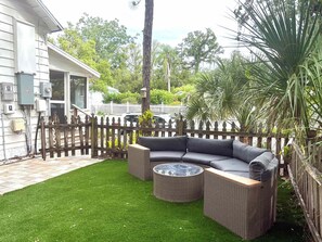 Eine Terrasse mit einem Ecksofa und einem runden Tisch, umgeben von einem Holzzaun und üppiger Bepflanzung.