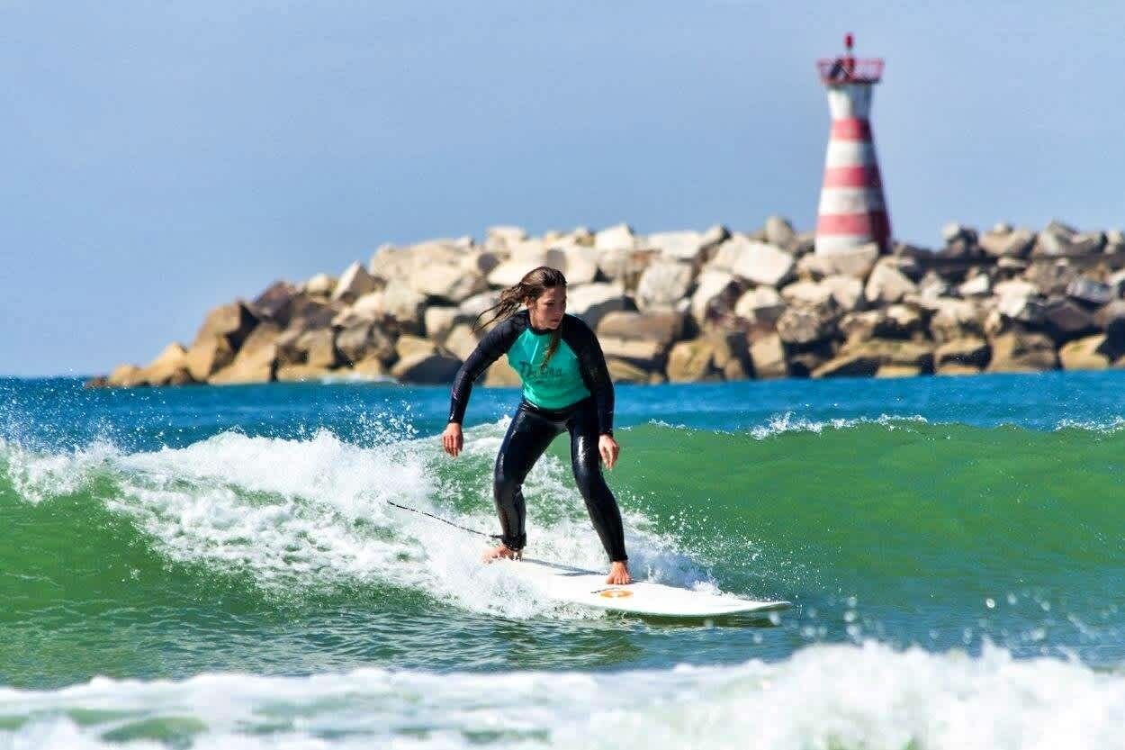 A person surfing on a wave with a rocky shoreline and a lighthouse in the background.