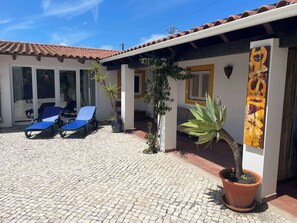Ein Gebäude mit einem Ziegeldach, einer überdachten Terrasse mit blauen Liegestühlen, Blumentöpfen und einem großen Wandkunstwerk.