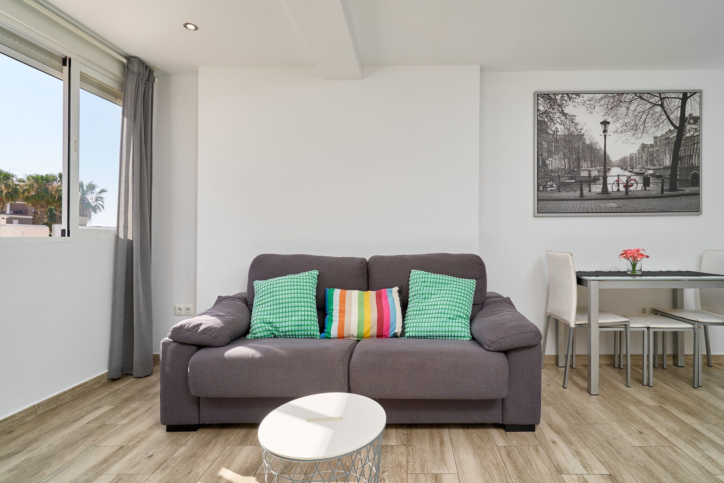 A modern living room with a grey sofa, a small white table, and a dining area with chairs and a table.