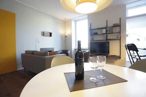 Ein modernes Wohnzimmer mit einer Couch, einem Esstisch mit Flasche und Gläsern, einem Fernseher und einem Bücherregal.
