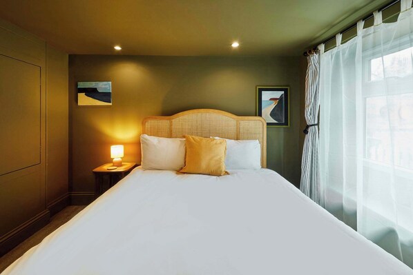 Una habitación de hotel con una cama grande, una mesita de noche con lámpara y una ventana con cortinas traslúcidas.