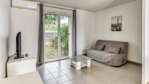 Un saló amb un sofà gris, una taula petita blanca, un televisor sobre un suport i una porta corredissa de vidre que dona a un jardí.