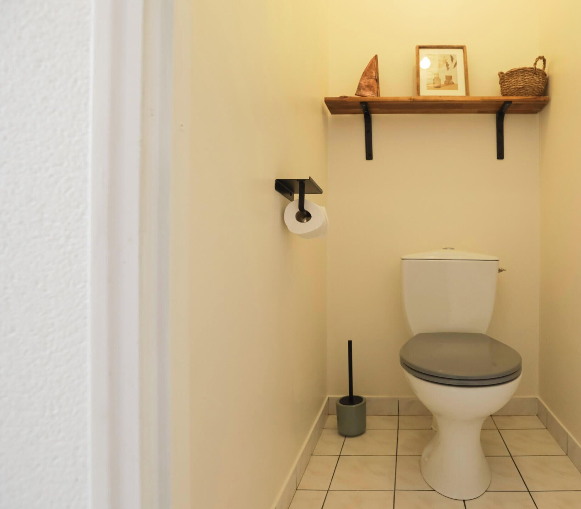 A bathroom with a toilet, a toilet paper holder, a bidet, and a shelf with a basket and a framed picture.