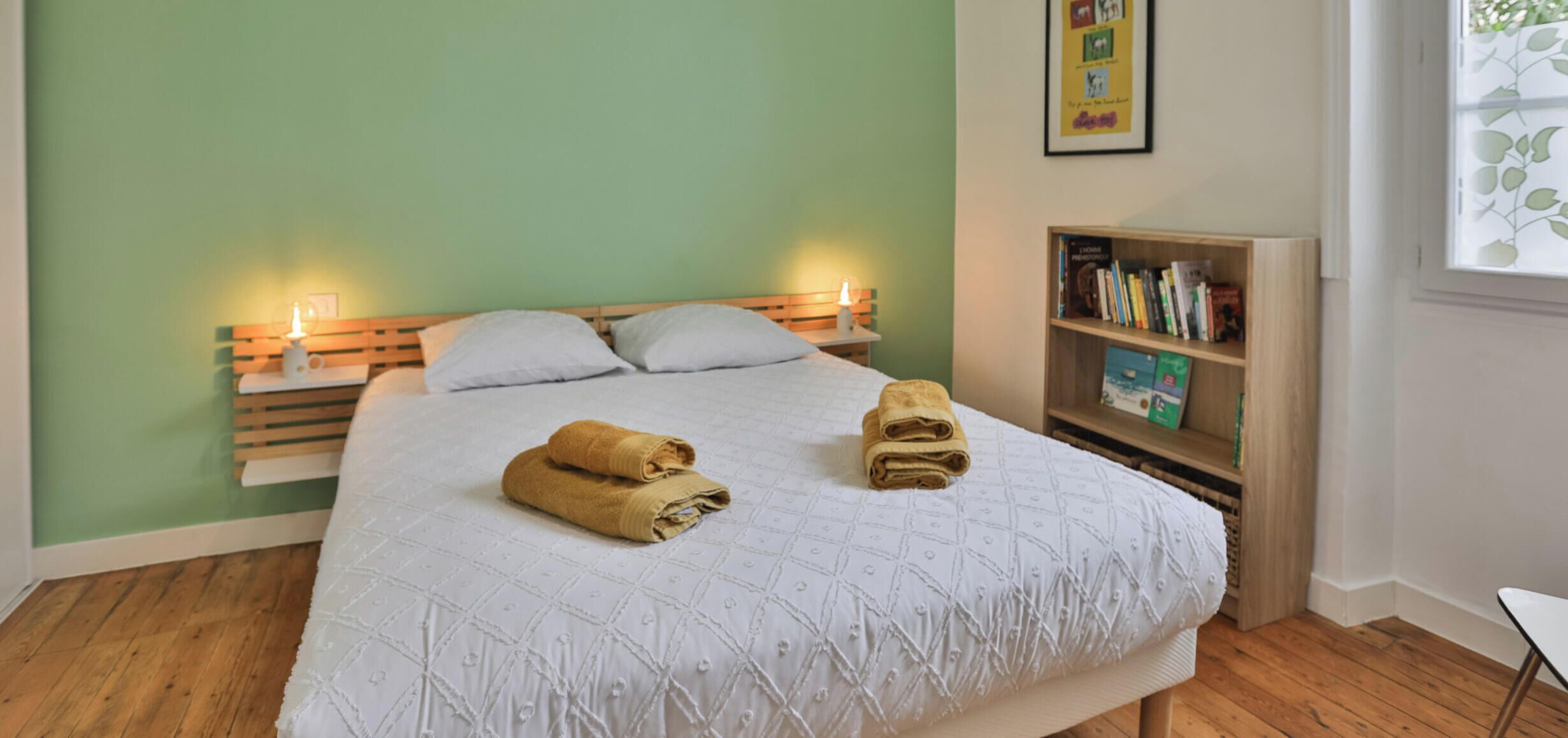 Una cama bien hecha con dos toallas enrolladas, una mesita de noche con una lámpara, una estantería y una ventana con una planta.