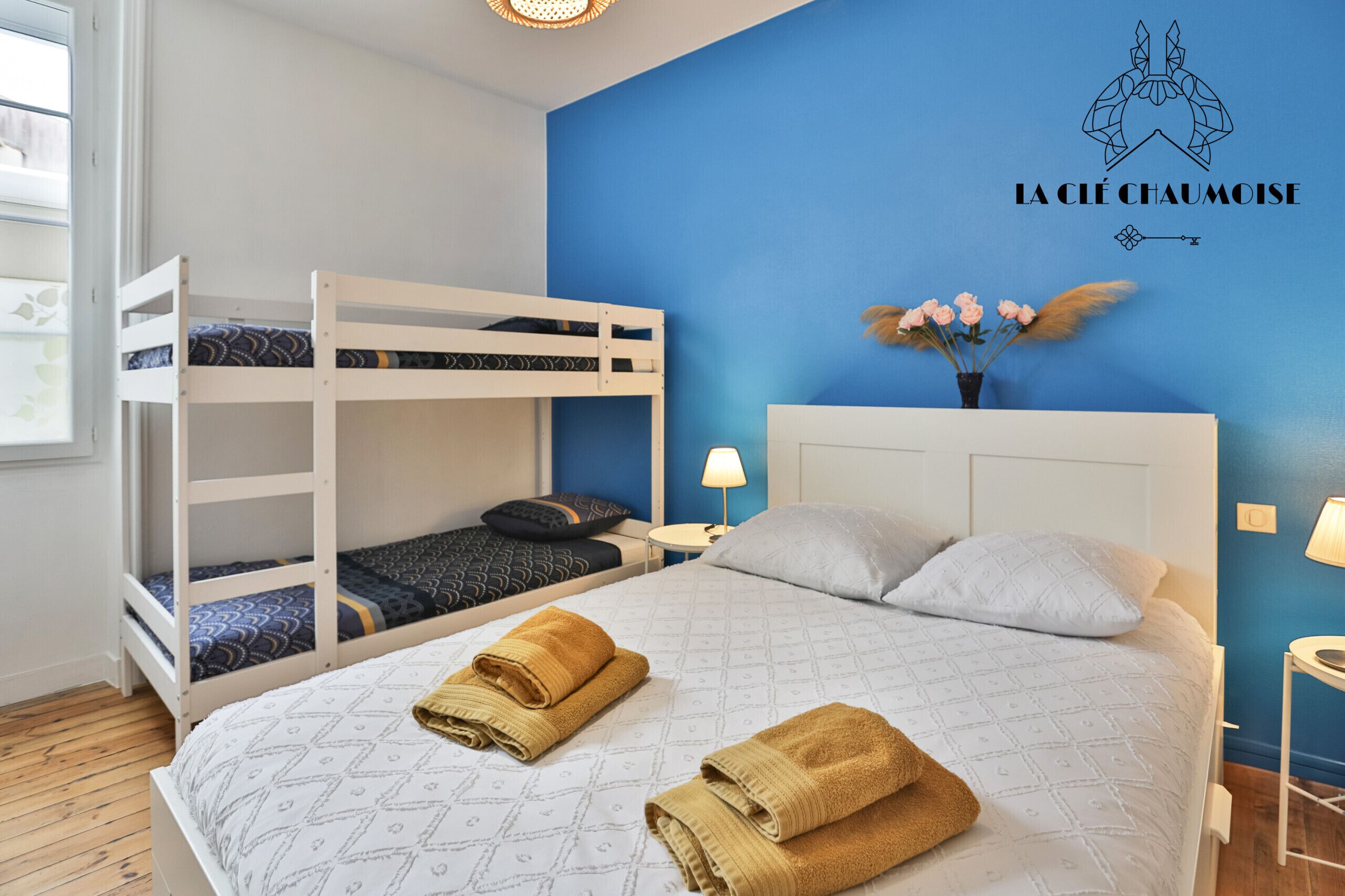 Een kamer met stapelbedden, een bed met handdoeken, een wandklok en een decoratie aan de muur.