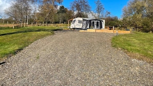 Een grindpad leidt naar een witte accommodatie in een camperstijl met een overdekte veranda, omgeven door bomen en een heldere lucht.