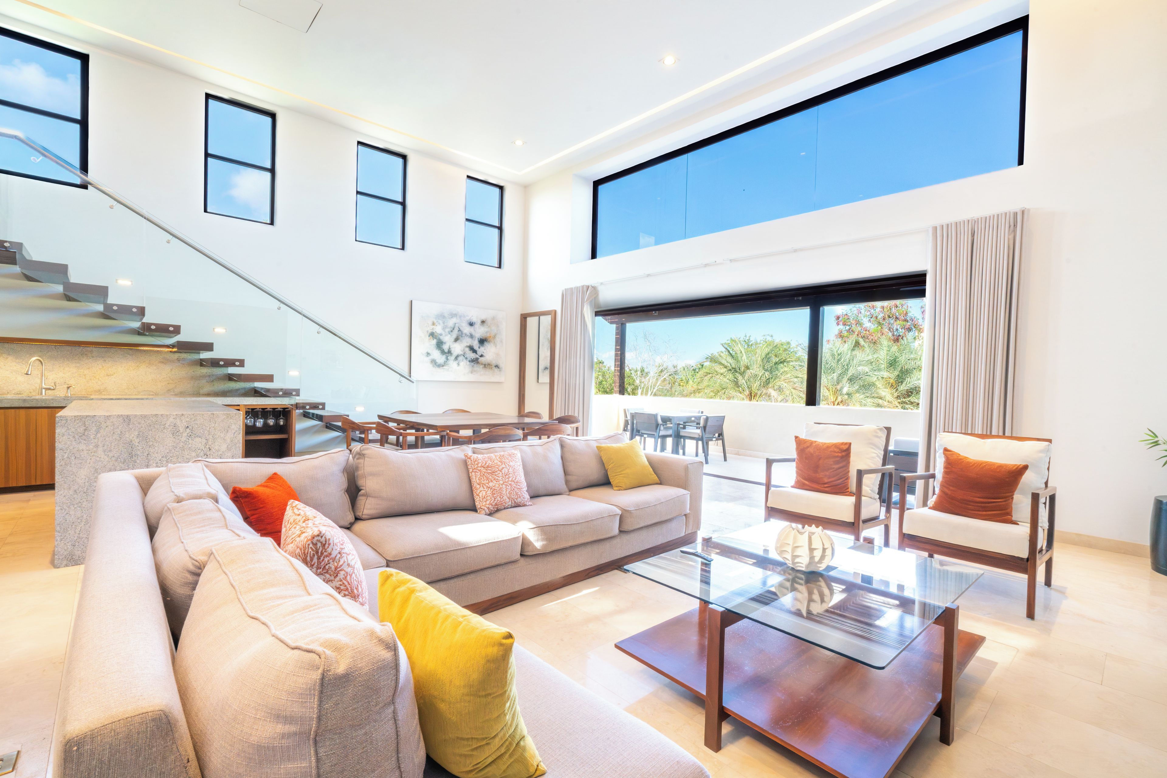 A spacious living area with a large sofa, coffee table, and seating arrangement. A staircase with glass railings leads to an upper level.