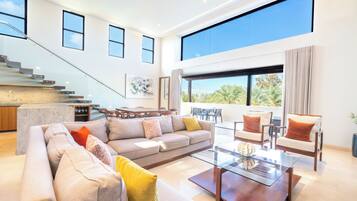 A spacious living area with a large sofa, coffee table, and seating arrangement. A staircase with glass railings leads to an upper level.