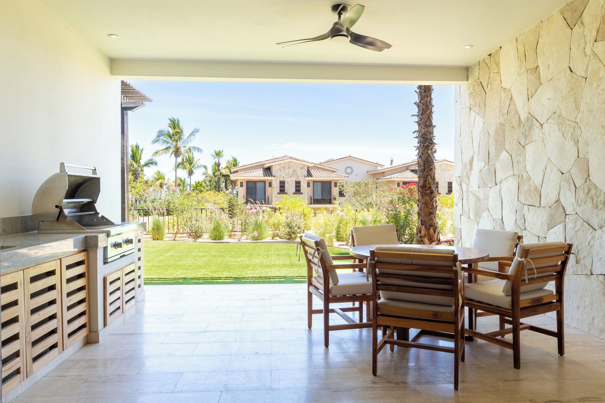 An outdoor kitchen with a grill, dining table, and chairs, overlooking a well-maintained lawn and buildings.