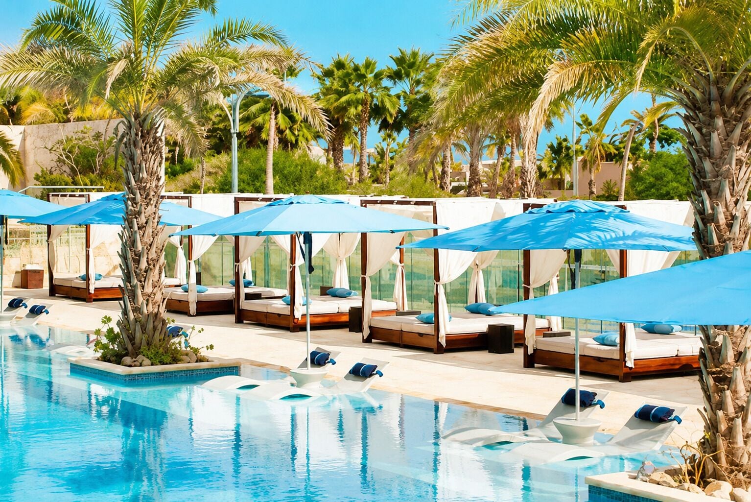 Ein Poolbereich mit mehreren Liegestühlen und Sonnenschirmen, umgeben von Palmen.