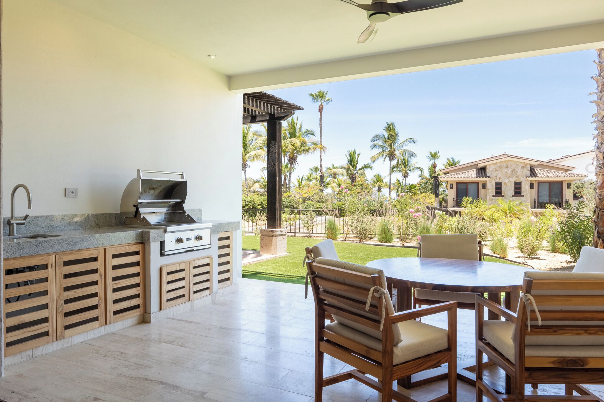 An outdoor kitchen with a grill, dining table, and chairs, overlooking a well-maintained garden with palm trees.