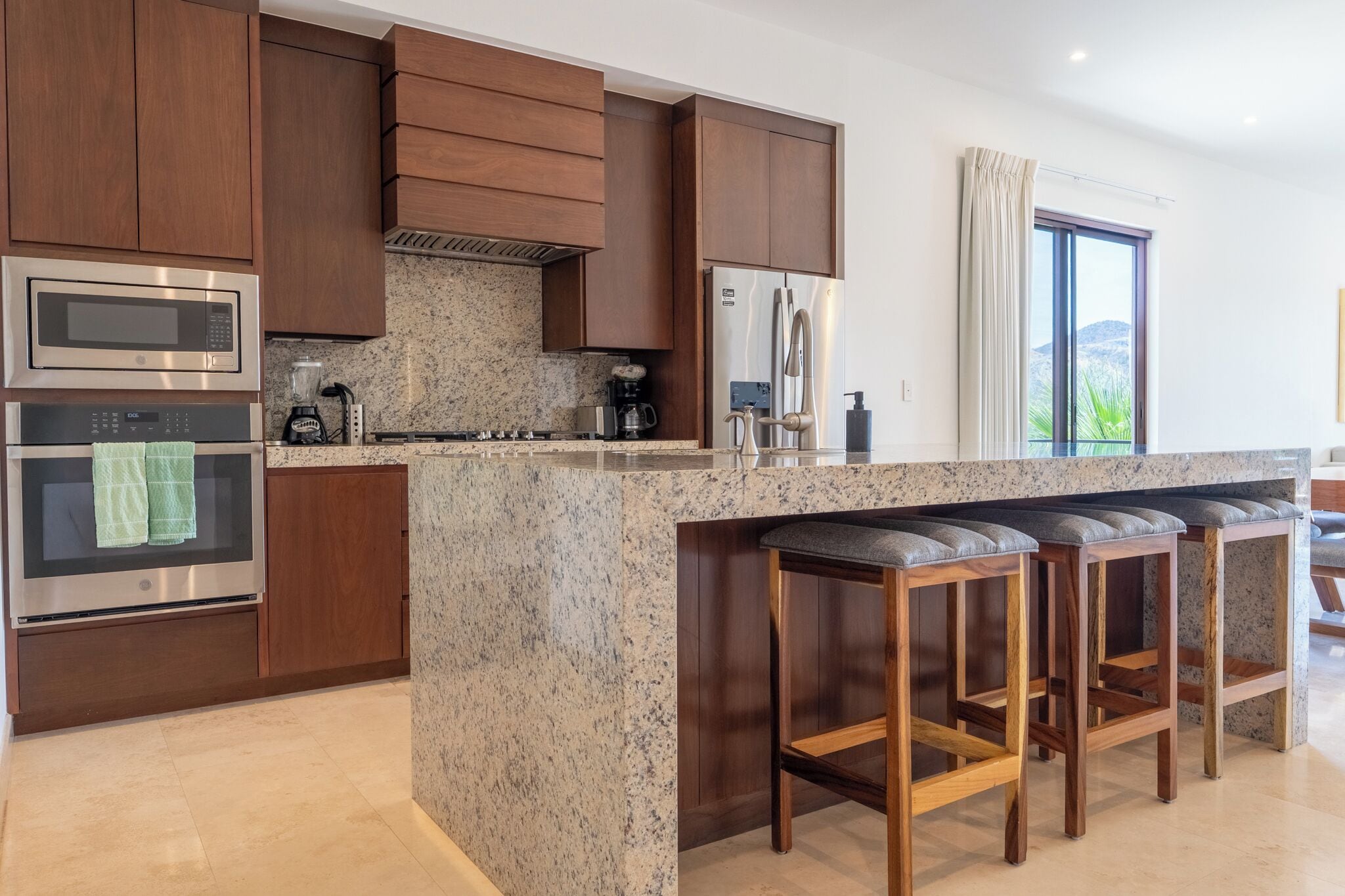 Una cocina moderna con gabinetes de madera, electrodomésticos de acero inoxidable, encimeras de granito y taburetes de bar.