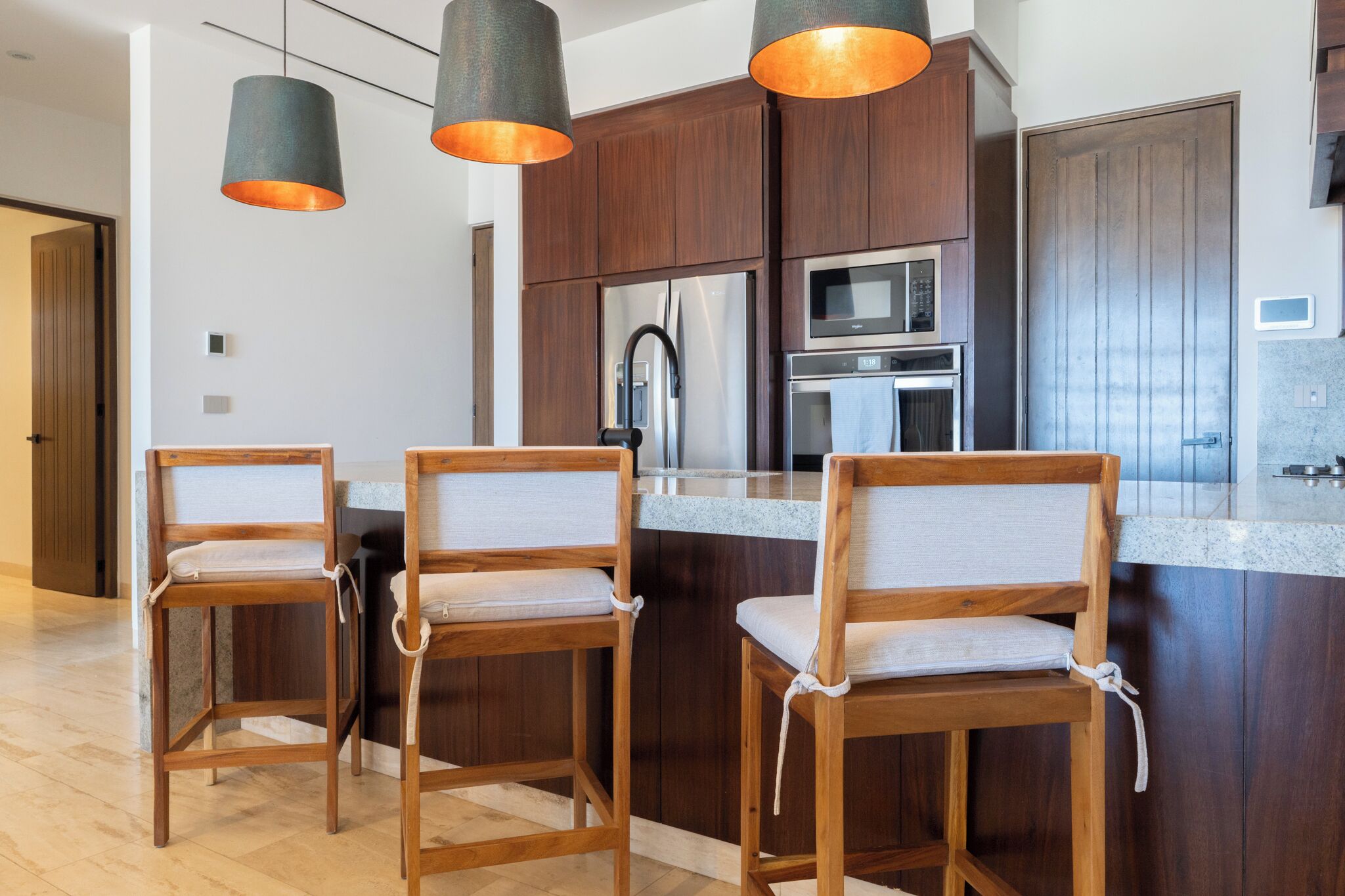 Een moderne keuken met donkerhouten kastjes, roestvrijstalen apparatuur en drie houten barkrukken met beklede zitplaatsen.