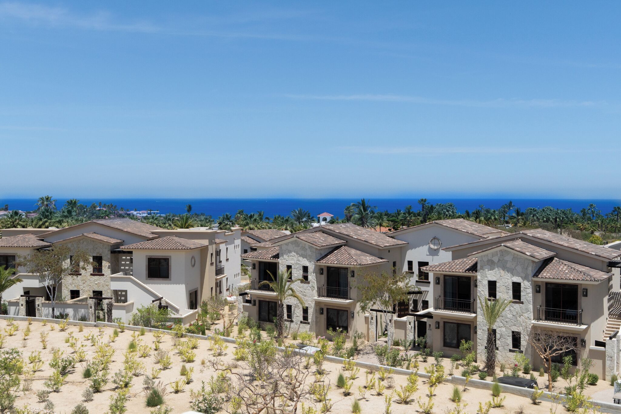 A residential area with multiple two-story houses, each with a balcony, surrounded by greenery and a clear view of the ocean.