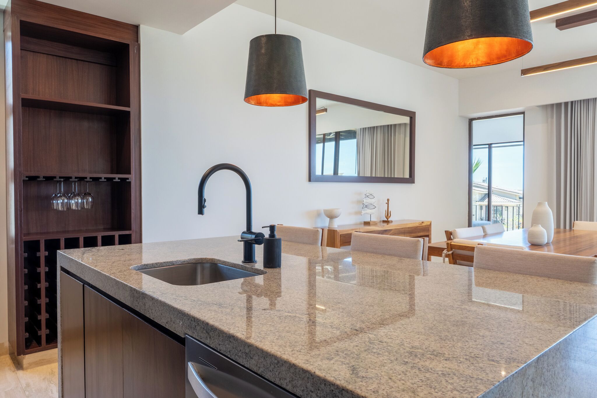 Una cocina moderna con encimeras de granito, una vinoteca empotrada, un grifo negro, luces colgantes, un comedor con mesa y sillas, y un espejo grande.