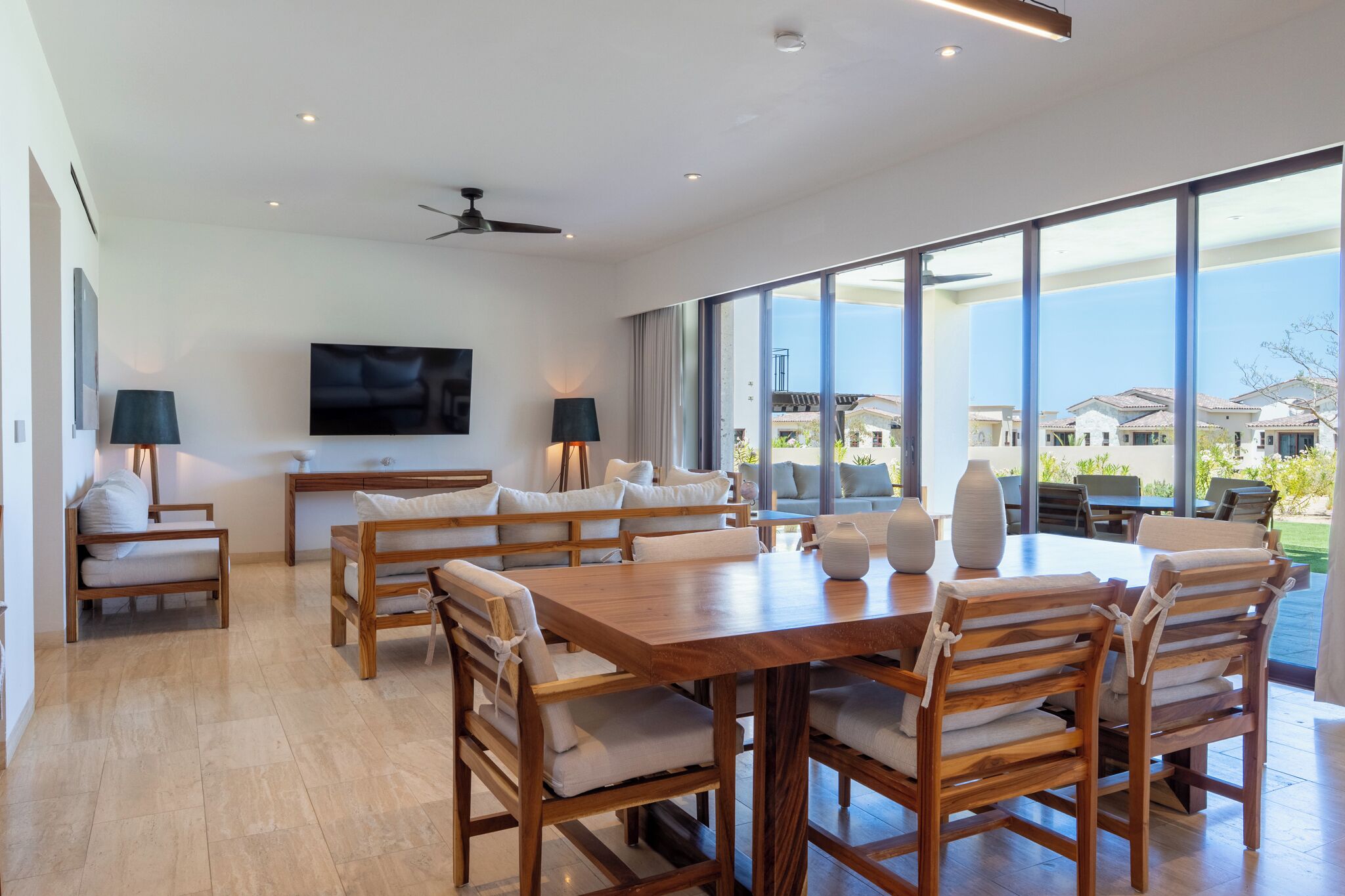 Un comedor espacioso con una gran mesa y numerosas sillas, un ventilador de techo y un amplio ventanal con vistas al vecindario.