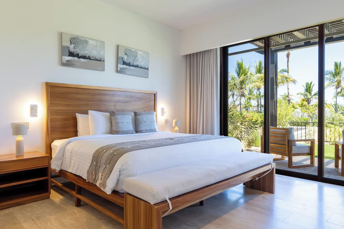 Un dormitorio moderno con una cama de madera, mesitas de noche y un amplio ventanal con vista a palmeras.