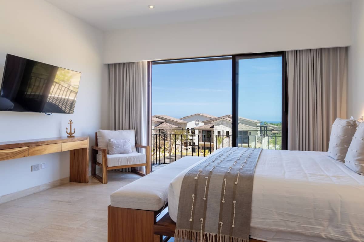 Un dormitorio con una cama grande, un escritorio, una silla, un televisor y un balcón con vistas a casas.