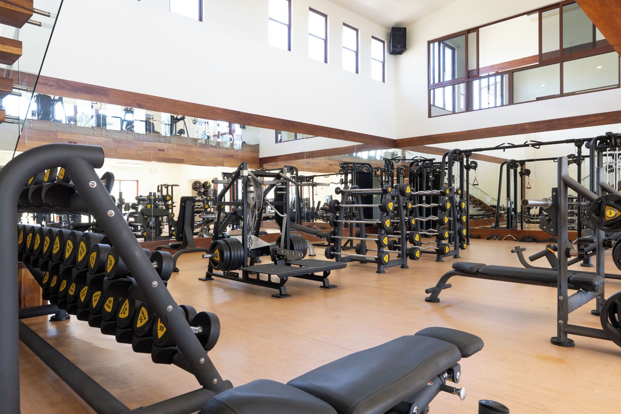 Ett gym med olika typer av träningsutrustning, trägolv och stora fönster.