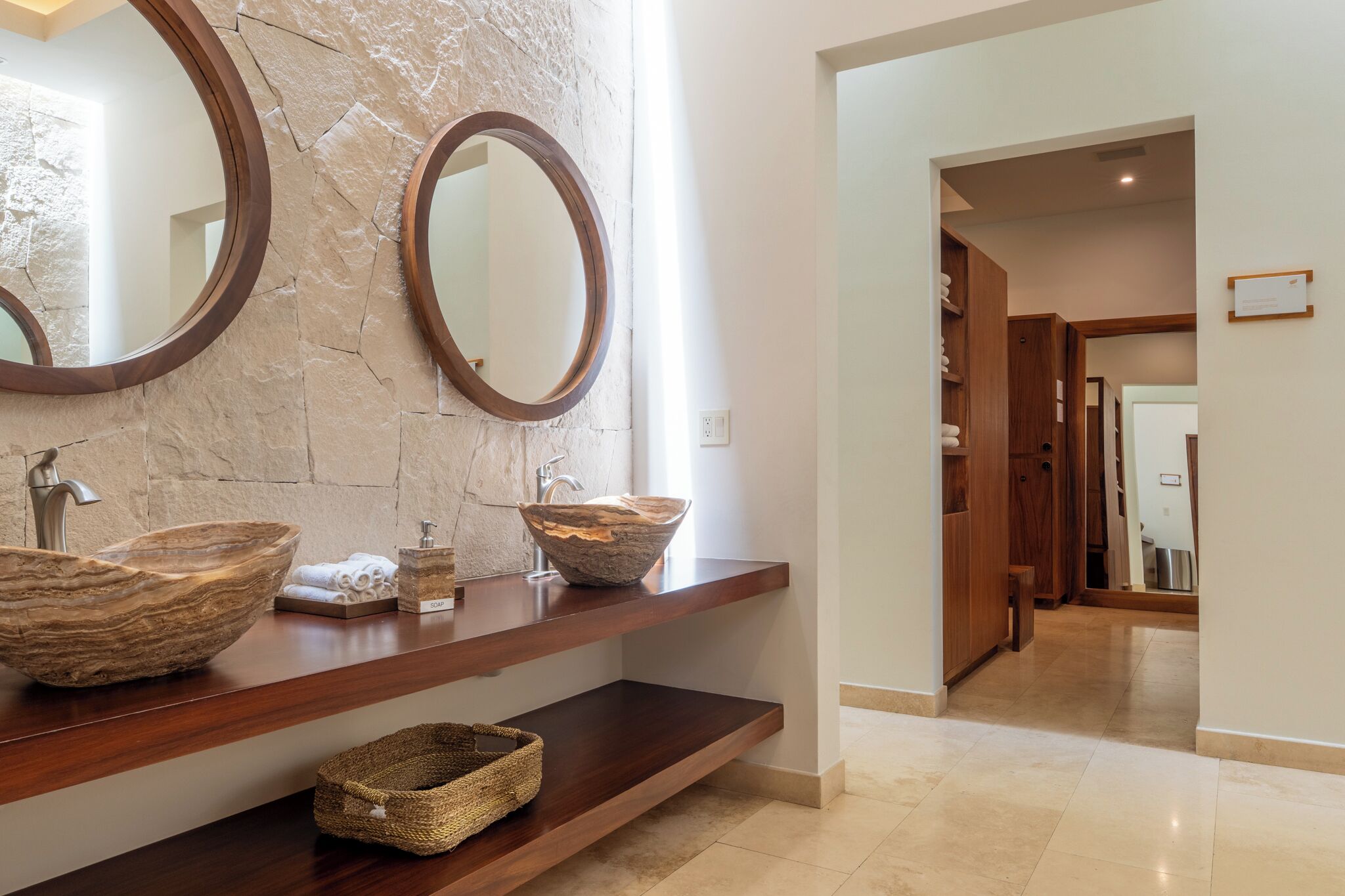 A bathroom with a stone wall, wooden vanity, two round mirrors, and a large bowl sink.