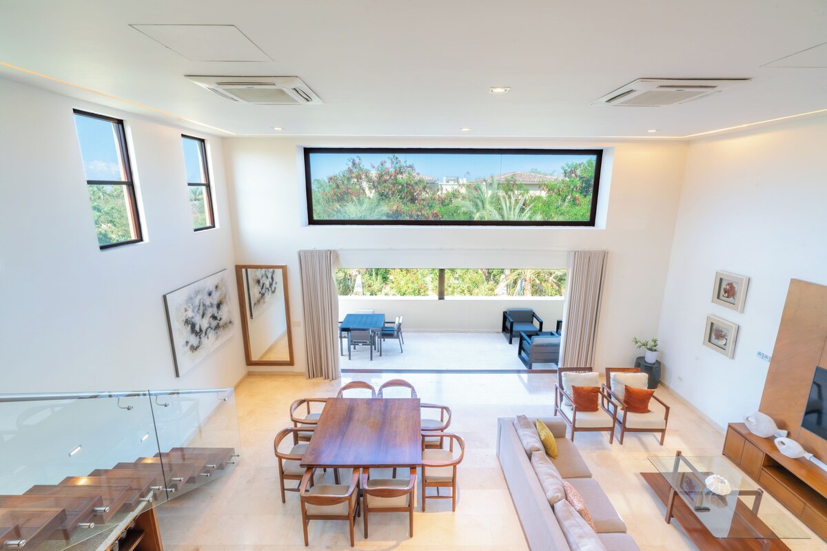 A modern living room with a dining area, a large flat-screen TV mounted on the wall, and a staircase leading to an upper level.
