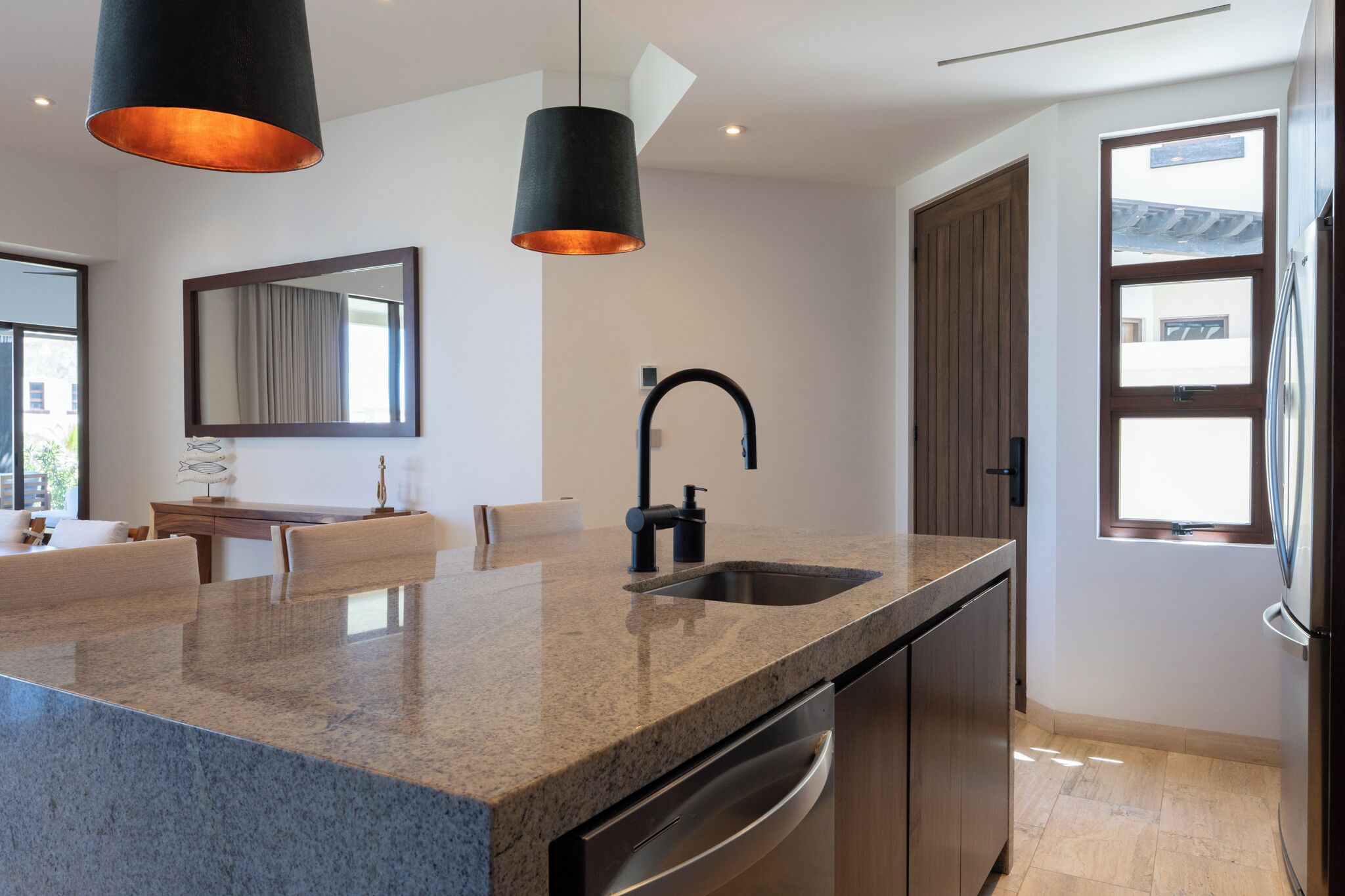 A modern kitchen with granite countertops, a built-in dishwasher, pendant lighting, a large mirror, and a window with a view.