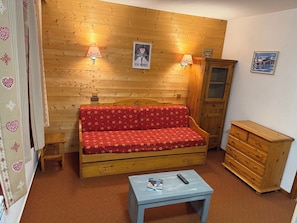 A cozy room with a wooden sofa, two wall-mounted lamps, a small table with a remote control, and a wooden cabinet.
