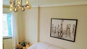 Een moderne woonkamer met een witte hoekbank, een ronde salontafel met eten erop, een kroonluchter en een ingelijst schilderij aan de muur.