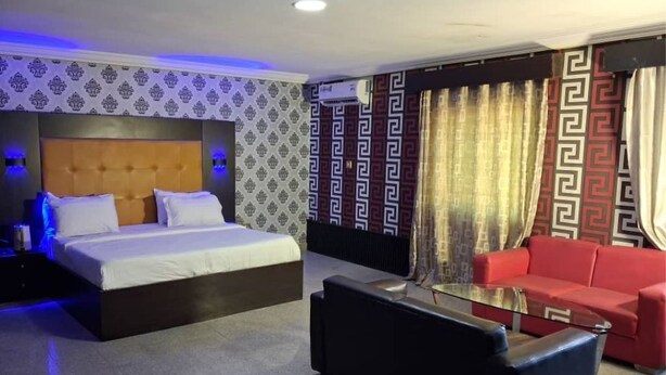Una habitación de hotel con una cama, un sofá rojo, una mesa de centro de vidrio, cortinas estampadas y un cabecero con una lámpara.