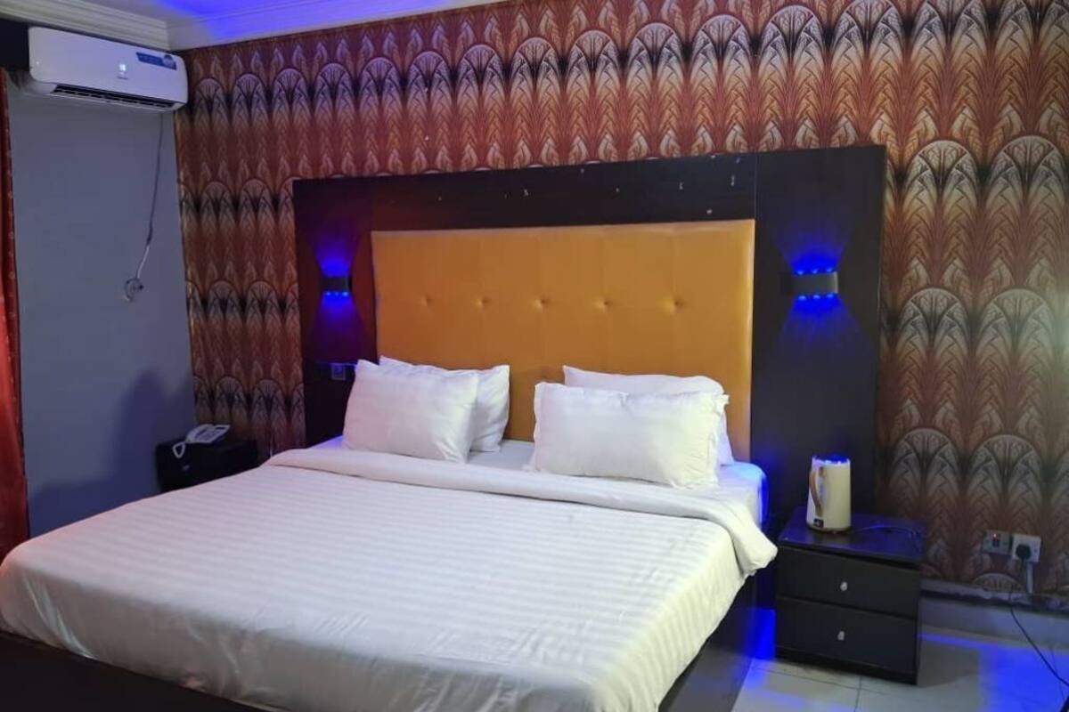 A hotel room with a large bed, a headboard with built-in lighting, a bedside table with a lamp, and patterned wallpaper.