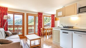 A modern kitchen with a microwave, dishwasher, and wooden cabinets. There is a dining area with a table and chairs, and a sofa in the living room. Large windows offer a view of the outdoors. - Stay for 5 Near Galibier Pass (Valloire)
