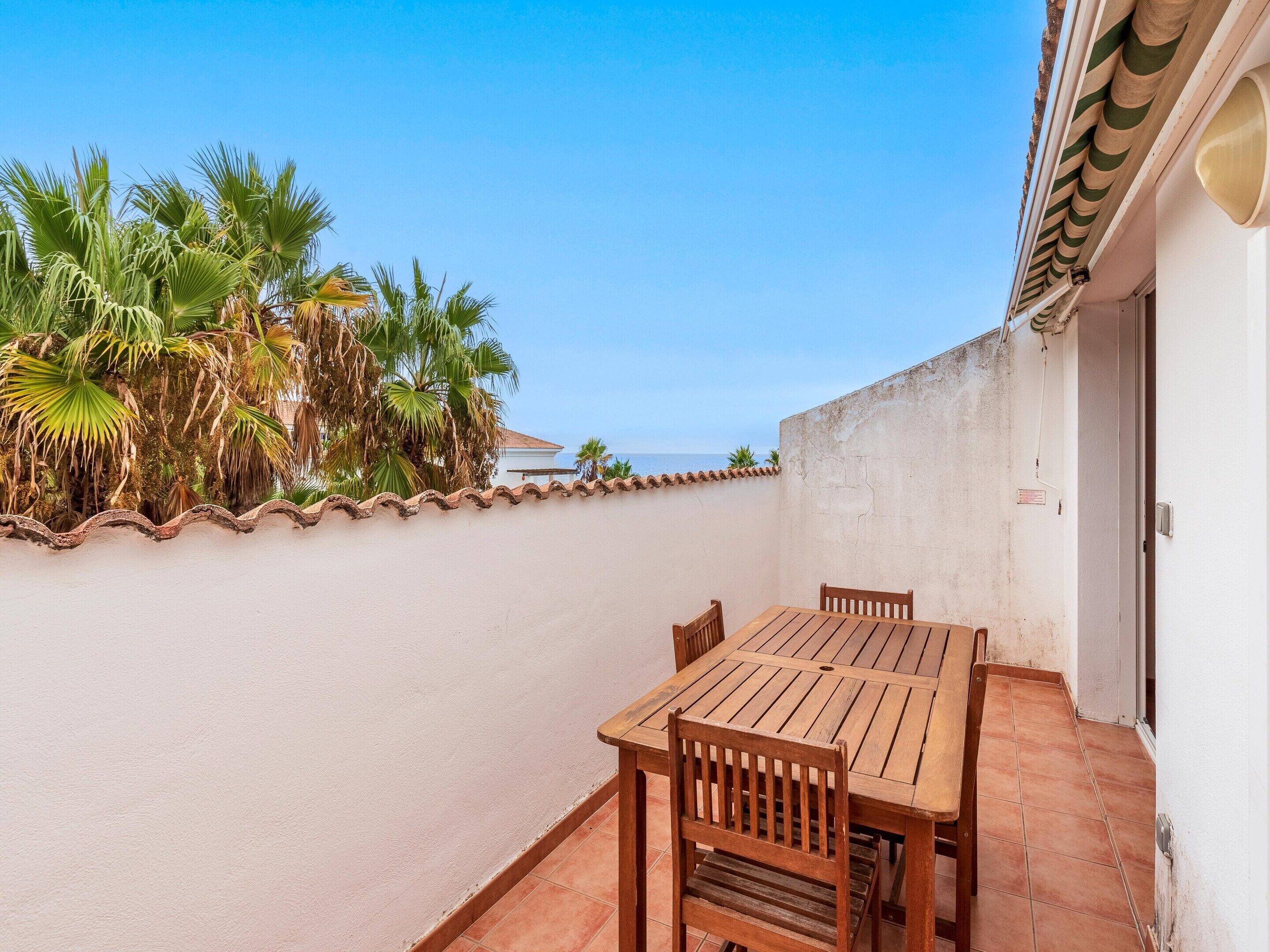 Un patio con una mesa y sillas de madera, una pared blanca y vistas a palmeras y al mar.