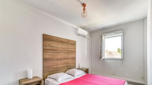 A bedroom with a wooden headboard, a bed with white bedding and a red blanket, a bedside table with a white object, and a window with a view of buildings. - Chic Coastal Pool View Apartment for 6 (Le Grau-Du-Roi)