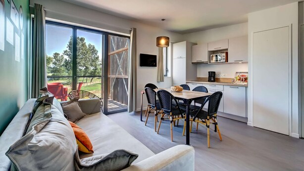 En moderne stue med sofa, spisebord, stole, et køkken med hårde hvidevarer og en stor skydedør til et udendørs område.