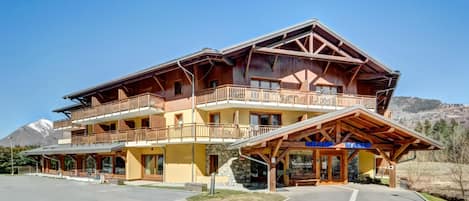 A multi-story hotel with balconies, a stone facade, and a mountain backdrop.